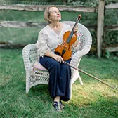 Kasia sitting In white wicker chair with violin